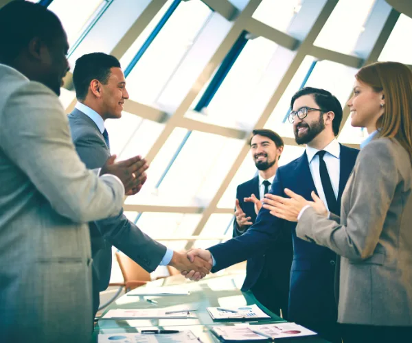 Action coach Building strategic partnerships, this image shows two business workers shaking hands with other employees clapping and smiling